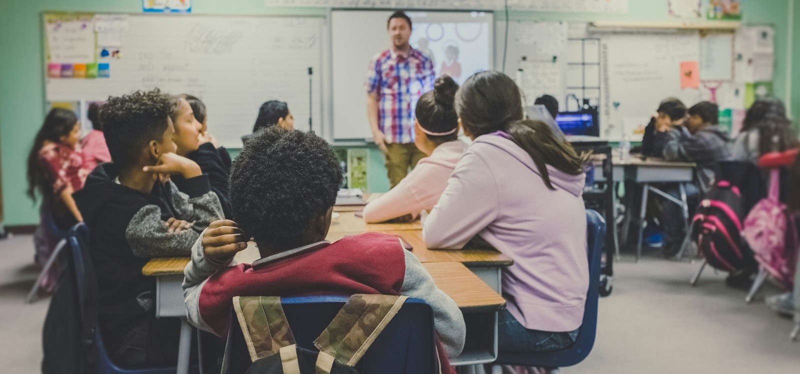 Students seated at desks facing a teacher who is standing at the front of a classroom.