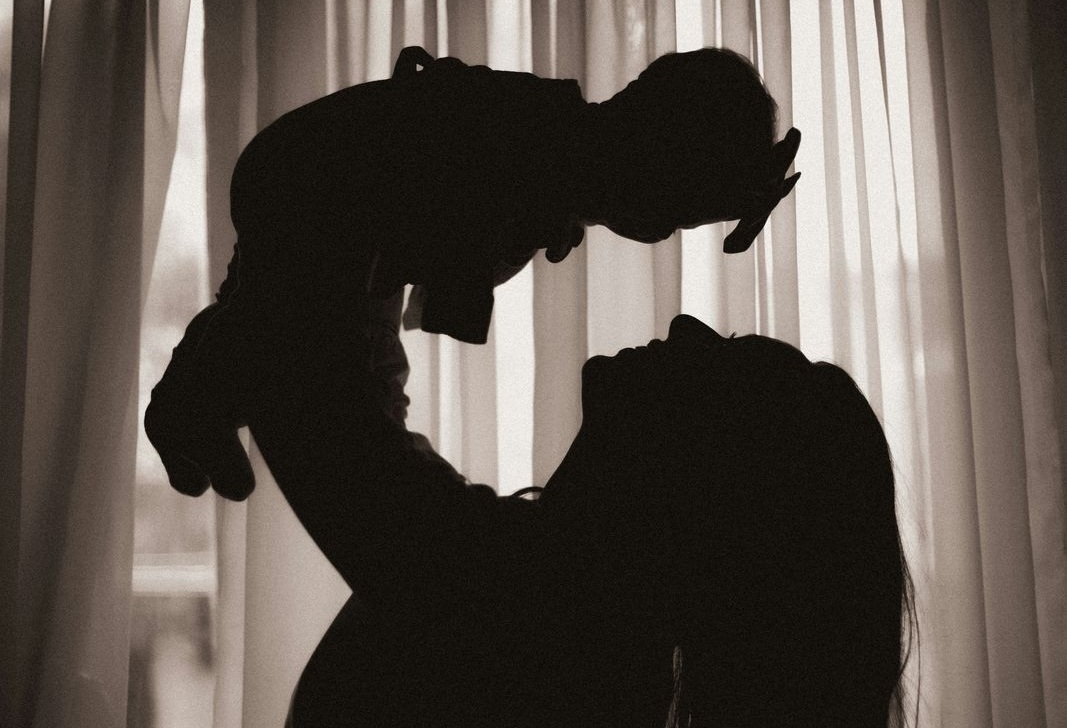 Silhouette of an adult lifting a baby in front of a window curtain.